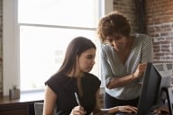 A woman in an office setting stands over a younger woman looking at a computer, helping her. 